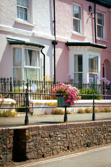 A view of a front garden in a residential area featuring a paved pathway bordered by a low brick wall and a black wrought iron fence. Behind the fence, there are two terraced houses with pastel-colore